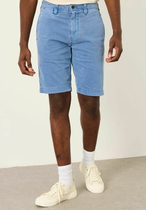 Mann trägt hellblaue Shorts, weiße Sportsocken und cremeweiße Sneakers, steht auf einem grauen Boden vor einer hellen Wand.