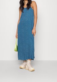 Modrá maxi šaty bez rukávů s tečkovaným vzorem, spárované s béžovými platformovými teniskami a malou zelenou kabelkou.