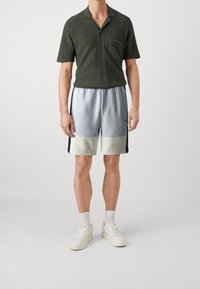 Chemise à manches courtes vert foncé avec des boutons et une poche, associée à un short de sport bleu clair et crème avec des bandes latérales. Baskets blanches.