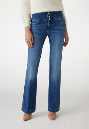 Femme portant un jean en denim bleu taille haute avec trois boutons à l'avant, un pull blanc et des talons pointus beiges, debout devant un fond uni.