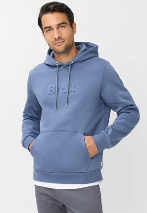Homme portant un sweat à capuche bleu avec poche avant et logo embossé "BRAX", les mains dans les poches, debout devant un fond clair uni.