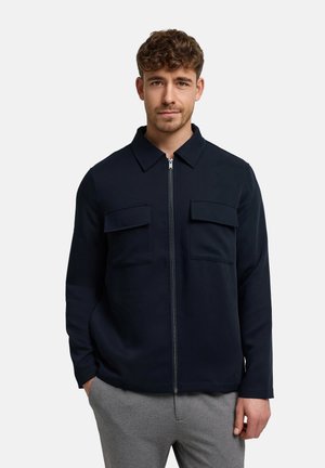 Mand med kort krøllet hår iført en marineblå jakke med lynlås og lommer med klap samt grå bukser, stående foran en ensfarvet lys baggrund.