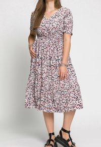 Robe fleurie à manches courtes, col en V, taille élastique, ornée de fleurs roses et rouges sur un fond blanc et noir. Portée avec des sandales noires.