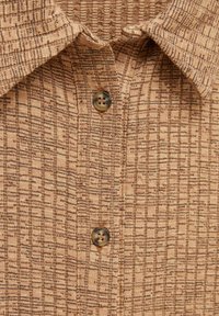 Col de chemise texturé marron avec deux boutons marron centrés sur un tissu à rayures verticales à motifs.
