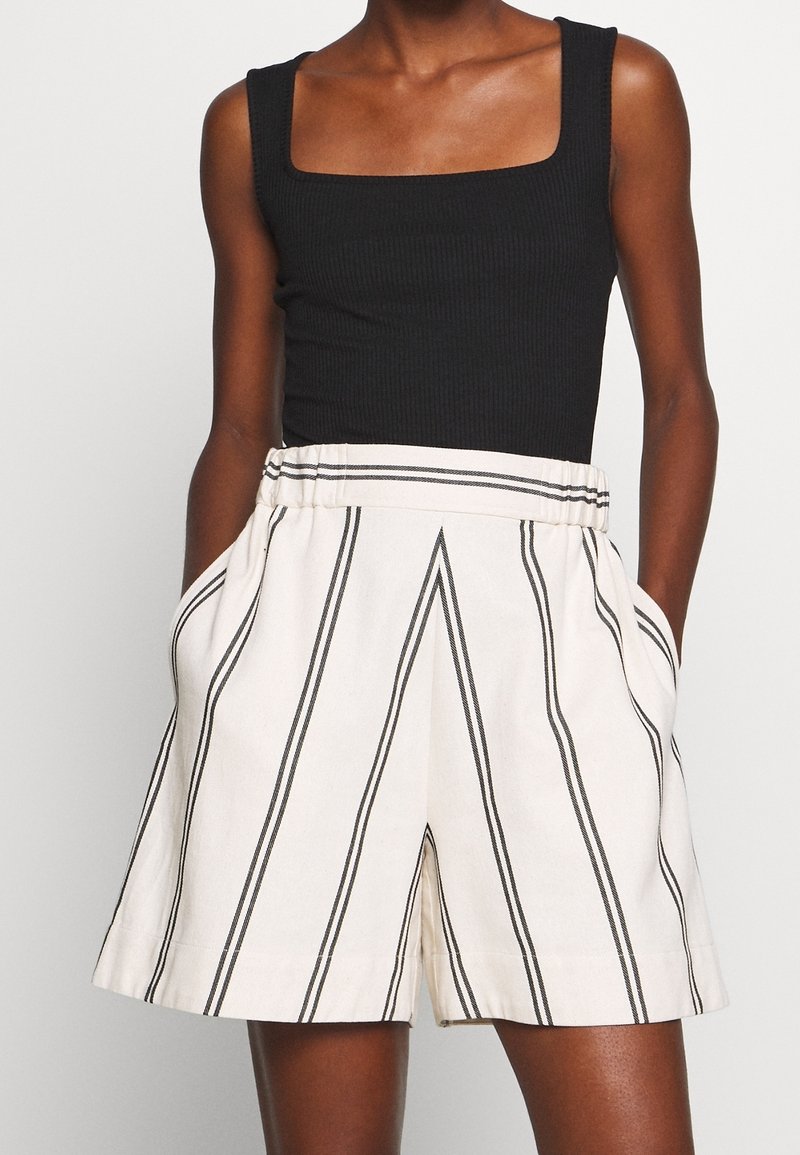 Personne portant un haut noir sans manches et un short blanc taille haute avec des rayures noires verticales et des poches latérales, debout devant un fond uni.