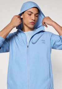 Ανοιχτό μπλε μπουφάν με φερμουάρ και κουκούλα, με λογότυπο Nike σε πιο σκούρο τόνο και ελαστικές μανσέτες. Υφή υφάσματος λείου τύπου.