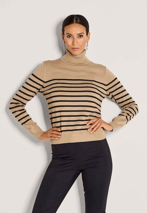 Frau mit dunklen Haaren, die einen beigefarbenen Rollkragenpullover mit schwarzen horizontalen Streifen und schwarze Hosen trägt, posiert mit den Händen in den Hüften.