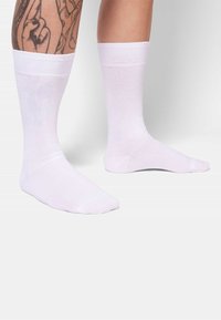 Weiße Baumwollsocken mit glatter Textur, gerippten Bündchen und knöchellang. Füße und ein tätowiertes Bein sind vor einem hellen Hintergrund sichtbar.