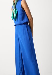 Haut bleu sans manches et pantalon à jambes larges en tissu lisse, agrémentés d'une écharpe verte à motifs nouée autour du cou, mettant en valeur des couleurs vives.