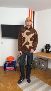 Chaqueta de forro polar marrón y topo con cremallera y patrón abstracto, combinada con jeans oscuros y zapatillas negras. Entorno interior con decoración mínima.