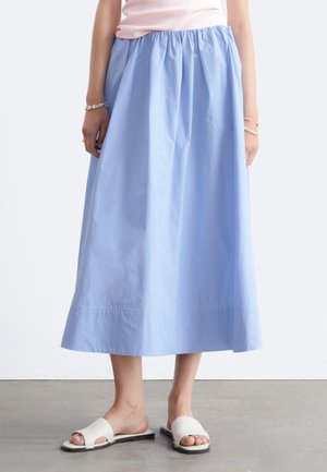 Femme portant une jupe midi bleu clair, un haut rose pâle, des sandales blanches plates à bout ouvert et des bracelets argentés debout sur un sol gris.