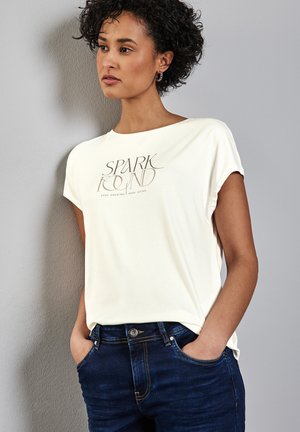 Un t-shirt léger à manches courtes de couleur crème présente un design graphique avec le texte "SPARK" en marron. Associé à un jean en denim bleu foncé, les mains dans les poches.