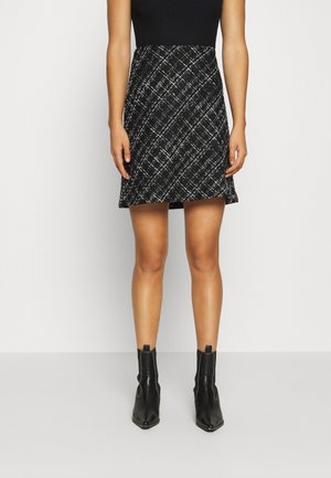 Femme portant une jupe à carreaux noir et blanc, un haut noir sans manches et des bottines noires, debout sur un sol gris clair.
