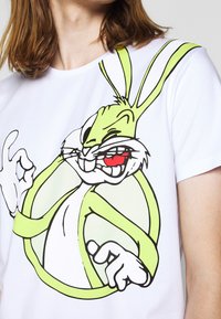 Bílý bavlněný tričko s grafikou kresleného králíka v zelené a černé barvě, s červenými akcenty a kulatým designem za ním.