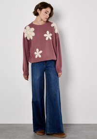 Helles rosa Sweatshirt mit großen weißen Blumenapplikationen, kombiniert mit weit geschnittenen blauen Denim-Jeans und braunen Schuhen. Gemütliches Freizeitoutfit.