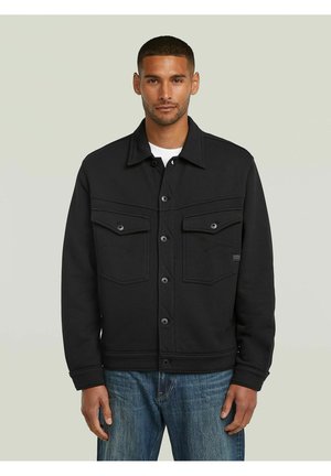 Homme portant une veste noire boutonnée avec deux poches poitrine et un jean bleu, debout devant un fond gris clair uni.