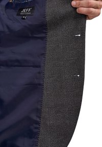 Interior de chaqueta entallada en negro y gris con un forro brillante en azul marino. Presenta costuras naranjas y detalles de sastrería visibles, incluidos los bordes sin rematar.