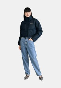 Giacca blu navy cropped e imbottita con colletto alto, caratterizzata da un logo sul petto; abbinata a jeans chiari a gamba larga e scarpe nere a punta.