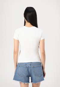 Hvit t-skjorte med korte ermer og tettsittende design, kombinert med blå denimshorts med slitt kant og to baklommer. Vist fra baksiden.
