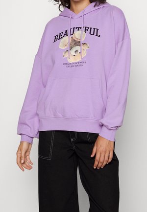 Person trägt einen lavendelfarbenen Kapuzenpullover mit Teddybär-Grafik und dem Text "SCHÖN" sowie "TRÄUME FUNKTIONIEREN NICHT, ES SEI DENN, DU ARBEITEST", kombiniert mit schwarzen Hosen.