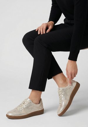 Persoon met zwarte broek en trui zit en raakt een metallic beige sneaker met bruine zool aan aan de rechtervoet.
