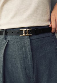 Cintura in pelle nera con fibbia metallica dorata che mostra un design a doppio anello, indossata sopra pantaloni grigi a trama con dettagli plissettati.
