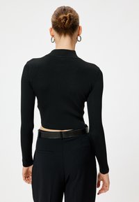 Top corto nero a coste con maniche lunghe, caratterizzato da un collo alto. Indossato con pantaloni neri a vita alta e una sottile cintura nera.