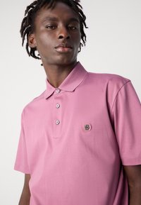 Gaišā rozā krāsā īsām piedurknēm polo krekls, izgatavots no gludas auduma, ar divu pogu kakla ailīti un logo uzlīmi kreisajā krūtīs.