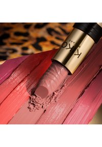 Lipstick tube with gold casing applies mauve shade over streaks of coral, red, mauve, and pink lipstick on a surface.