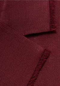 Gros plan d’un tissu tissé bordeaux plié avec une texture fine et des bords frangés sur deux sections chevauchantes.
