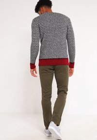 Schwarz-weißer Musterpullover mit rotem Besatz, kombiniert mit olivgrünen Hosen und weißen Sneakers. Die Textur wirkt gestrickt.