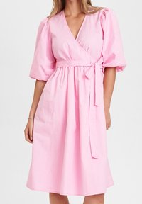 Kvinna i en knälång, ljusrosa omlottklänning med puffsleeves och ett knytbälte i midjan, stående mot en neutral bakgrund.