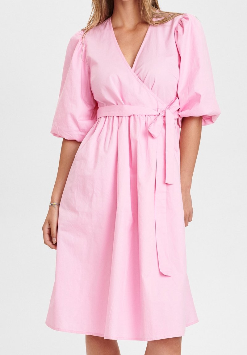 Kvinna i en knälång, ljusrosa omlottklänning med puffsleeves och ett knytbälte i midjan, stående mot en neutral bakgrund.