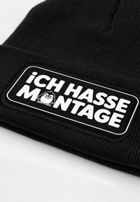 Black knit beanie with ribbed texture, featuring a rectangular white patch. Text reads "Ich hasse Montage" with a cartoon penguin graphic.