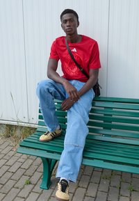 Red Adidas t-shirt with FC Bayern Munich logo, light blue baggy jeans, and beige sneakers with black stripes, seated on a green bench.