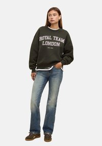 Donna che indossa un felpa verde oliva con la scritta "ROYAL TEAM LONDON", jeans blu chiaro e scarpe da ginnastica verdi, in piedi con una mano in tasca.
