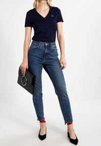 Granatowa koszulka z dekoltem w serek w zestawie z niebieskimi, wysokogatunkowymi dżinsami, z czerwonym akcentem na mankietach, czarnymi szpilkami oraz czarną kopertówką z okrągłym uchwytem.