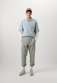 Chemise bleue à manches courtes en coton texturé, pantalon de jogging gris avec des poignets élastiques, casquette beige et baskets gris-vert.