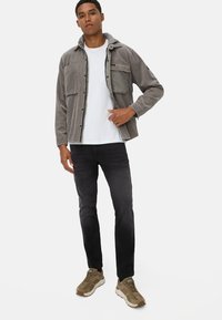 Graues Cordhemd mit Knopfleiste und Kapuze, zwei Brusttaschen, über einem weißen T-Shirt getragen, kombiniert mit schwarzen Jeans und braunen Sneakers.