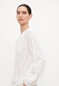 Hvit langermet bluse med blonder, rund halsutskjæring, samlede ermer og myk tekstur. Har diskrete broderte detaljer.