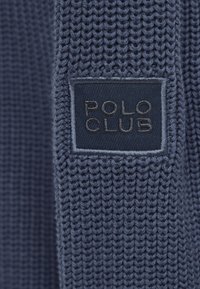 Gestrickter marineblauer Stoff mit einer strukturierten Webart und einem rechteckigen schwarzen Etikett mit geprägten Buchstaben "POLO CLUB" auf der Oberfläche.