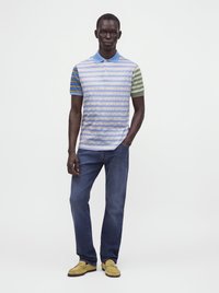 Homme debout une main dans la poche portant un polo à rayures multiples, un jean bleu et des mocassins jaunes sur un fond blanc uni.