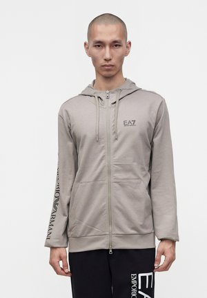 Homme portant un sweat à capuche gris clair zippé avec des poches frontales et un texte noir sur la manche gauche et la poitrine, associé à un pantalon noir avec un texte blanc.