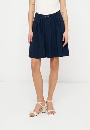 Femme portant une jupe plissée bleu marine avec une boucle décorative à la taille, un haut blanc et des sandales à talons blocs beiges à brides.