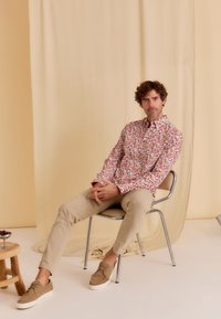 Langarmshirt met een rood en groen bladerenpatroon, gecombineerd met beige tapered broek en lichtbruine schoenen, zittend op een stoel.