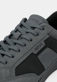 Baskets en daim gris avec accents noirs texturés, design à lacets et logo de marque bien visible. Semelle extérieure en caoutchouc lisse.