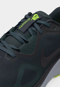 Κοντινό πλάνο σε μαύρο αθλητικό παπούτσι με μπλε κορδόνια, μαύρο λογότυπο Nike και φωτεινή πράσινη ετικέτα που γράφει "RUN Seltucture 26."