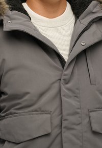 Grijze winterjas met een getextureerde kraag, twee zakken aan de voorkant en een ritssluiting. De stof lijkt glad en geïsoleerd.