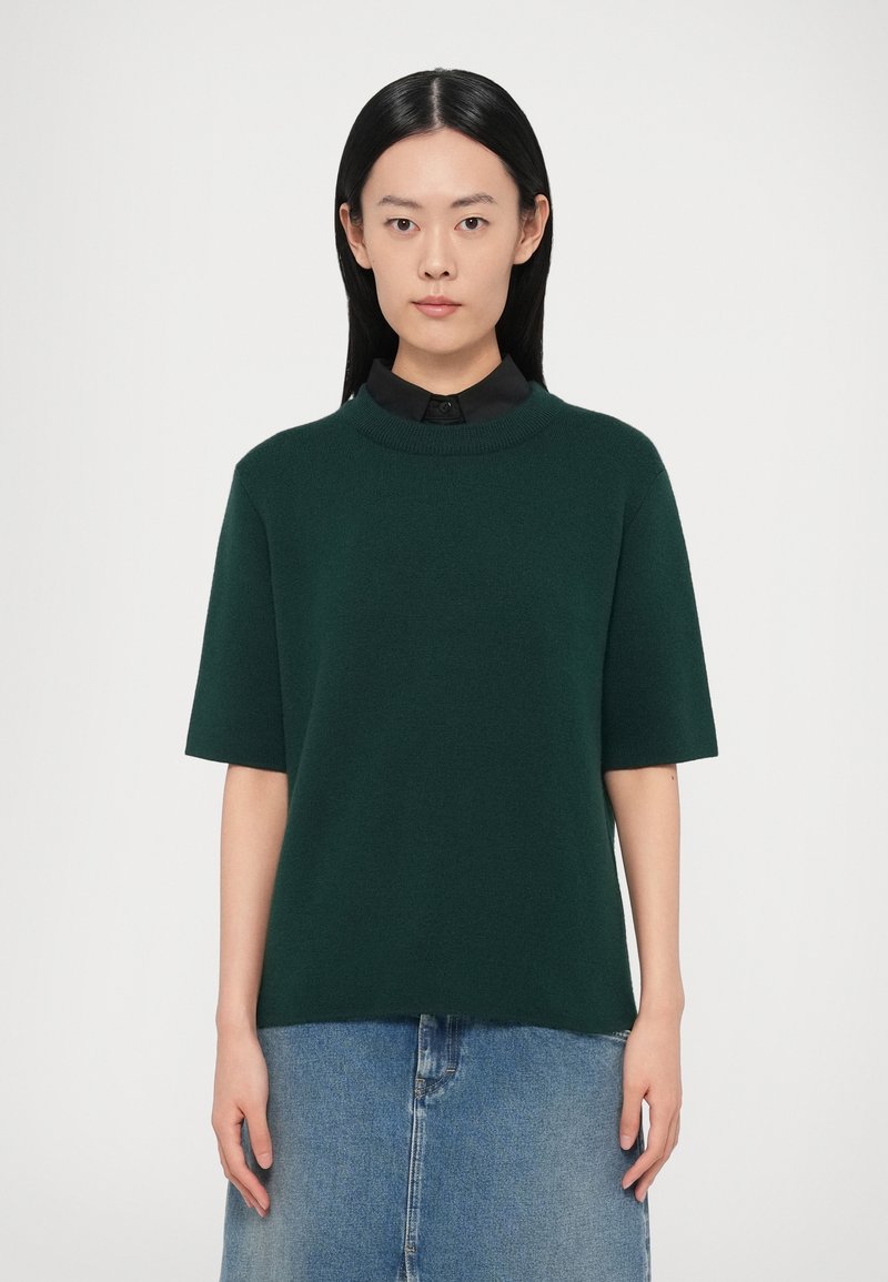 Tumši zaļš adīts džemperis ar īsām piedurknēm un augstu apkakli, kopā ar džinsa svārkiem. Vienmērīga tekstūra, brīvs piegriezums.