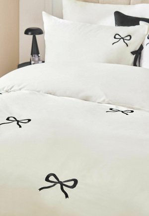 Weiße Bettwäsche mit schwarzen Schleifendekoren, weiße und schwarze Kissen sowie eine schwarze Nachttischlampe auf einem Nachttisch in einem Schlafzimmer.